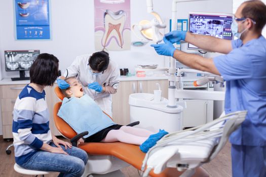 Kid patient in dental office getting teeth treatment sitting on dental chair wearing bib with mouth open. Dentistry specialist during child cavity consultation in stomatology office using modern technology.