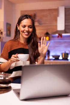 Young woman holding cup of tea using video call concept for online conference on laptop in festive kitchen at home. Caucasian adult enjoying chat with relatives on christmas eve night