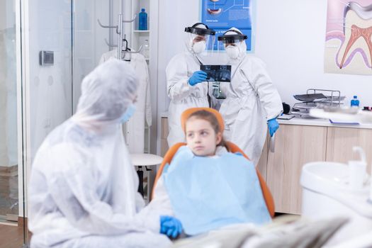 Dentist team analyzing kid radiography dressed in protection gear against coronavirus. Stomatologist during covid19 wearing ppe suit doing teeth procedure of child sitting on chair.