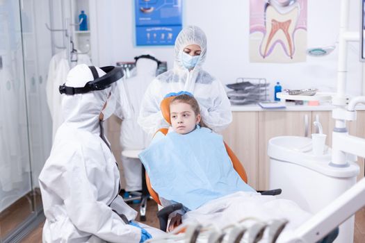 Dentist in coverall talking with kid before dentistiry examination with face mask. Stomatologist during covid19 wearing ppe suit doing teeth procedure of child sitting on chair.