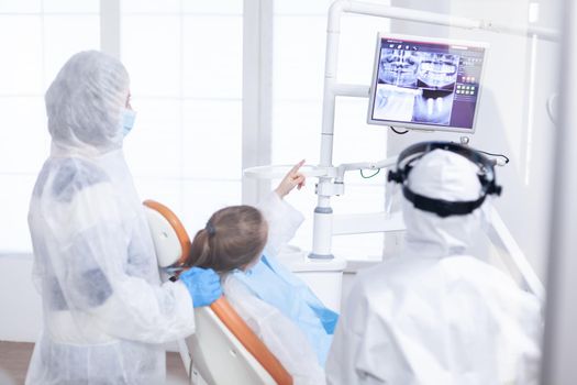 Little girl in ppe suit pointing at digital radiography during consultation. Stomatolog in protectie suit for coroanvirus as safety precaution looking at digital child teeth x-ray during consultation.