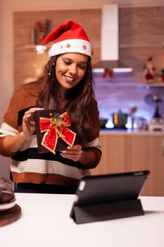 Woman with santa hat using video call conference on tablet in kitchen with ornaments, decorations and lights at home. Caucasian adult showing surprise gift with bow for christmas eve party