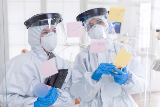 Dentists discussing about patient teeth dressed in ppe suit during covid-19. Medical team in stomatology office wearing coverall in dental office writing ideas on sticky notes.