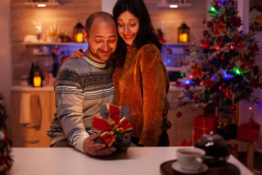 Happy married couple enjoying christmas holiday spending winter festive season together in xmas decorated kitchen. Husband holding secret gift celebrating x-mas wintertime