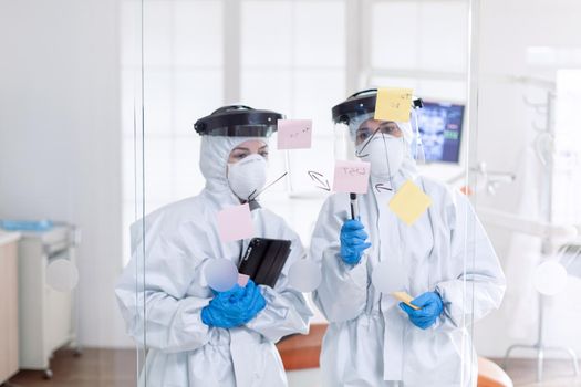 Professional dentists in ppe suit checking schedule during covid-19. Medical team in stomatology office wearing coverall in dental office writing ideas on sticky notes.