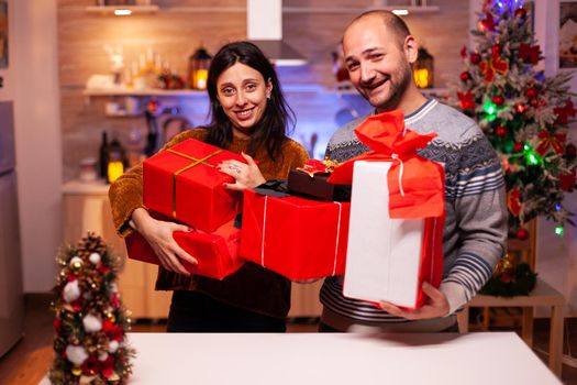 Happy married couple holding secret present gift with ribbon on it during christmas holiday standing in xmas decorated kitchen. Smiling family enjoying winter season celebrating christmastime together