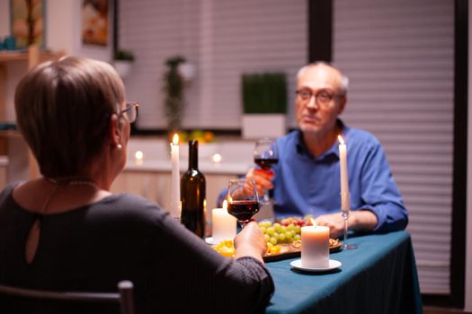 Woman having a romantic dinner with husband in kitchen in the evening holding glass of wine. Senior couple sitting at the table in dining room , talking, enjoying the meal, celebrating their anniversary in the dining room.