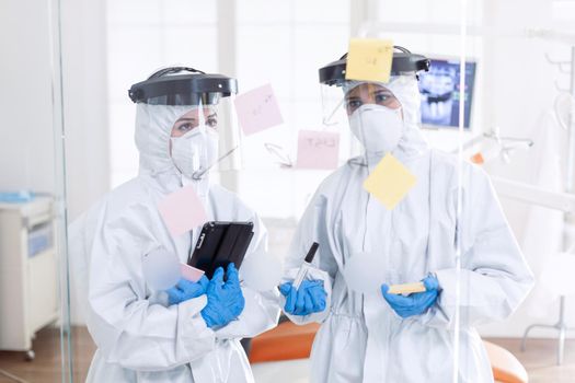 Dentist during coronavirus wearing ppe suit planing patient treatment. Medical team in stomatology office wearing coverall in dental office writing ideas on sticky notes.