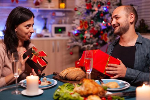 Happy married couple holding secret present with ribbon on it during christmas holiday sitting at in xmas table in decorated kitchen. Smiling family enjoying winter season celebrating christmastime