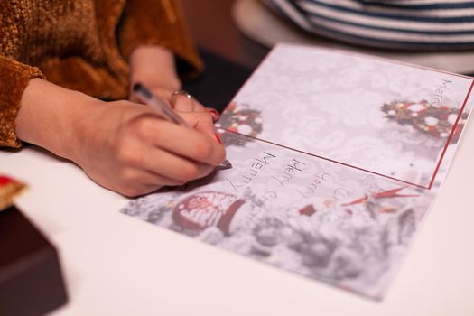 Closeup of woman hands writing xmas greeting card for family during christmas holiday standing in xmas decorated kitchen. Happy family enjoying spending winter season together