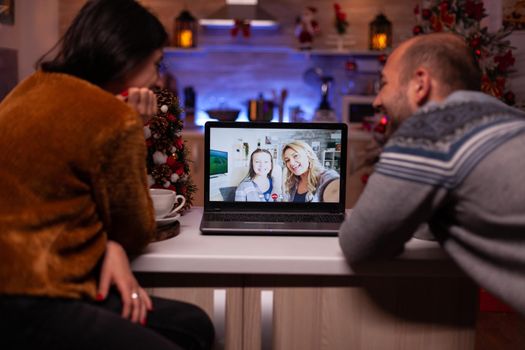Joyful family talking with remote friends during online videocall meeting on laptop computer celebrating christmastime together in xmas decorated kitchen. Happy couple enjoying winter season