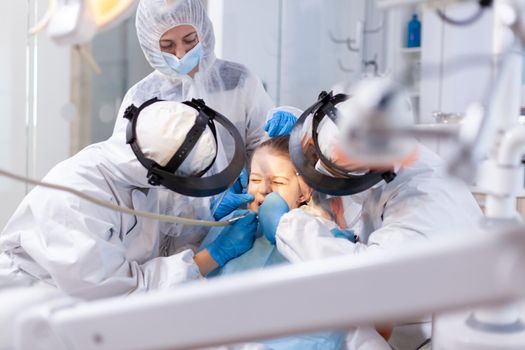 Little girl with painfull expresion in the course of dentistiry dental treatment dressed in ppe suit. Stomatology team wearing ppe suit during covid19 doing procedure on child teeth.