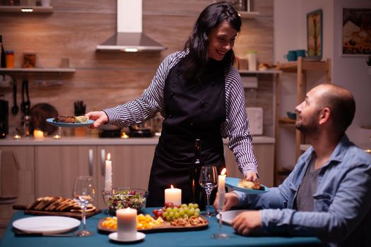 Wife surprising husband with dinner in dining room and celebrating with delicious food. Woman preparing festive dinner, cooking for his man romantic dinner, talking, sitting at table