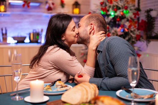 Happy couple kissing in xmas kitchen after marriage proposal enjoying christmas holiday sitting at dinning table. Couple enjoying spending time together during winter season