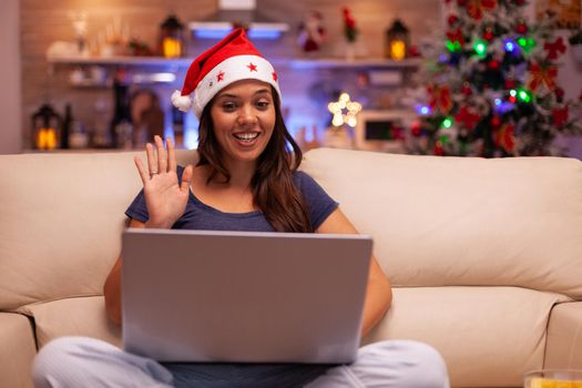 Girl greeting remote friend during online videocall conference meeting on laptop resting on couch in xmas decorated kitchen. Woman celebrating christmas holiday enjoying winter season