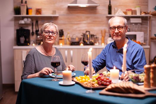 Joyful couple in kitchen looking at camera holding wine glasses looking at camera during dinner. Happy cheerful senior elderly couple dining together in the cozy kitchen, enjoying the meal.