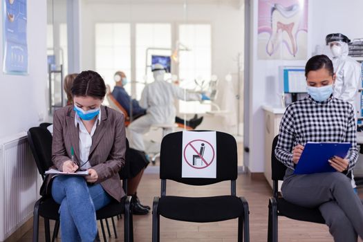 Patient with face protection mask writing on registration form in stomatomoly clinic, sitting in reception respecting social distance. Dentistiry doctor receptionist dressed in ppe suit.