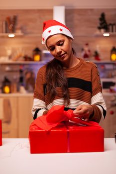 Adult with santa hat wrapping bow on present box in christmas decorated kitchen at home. Caucasian young woman preparing gift for friends and family at festive december celebration