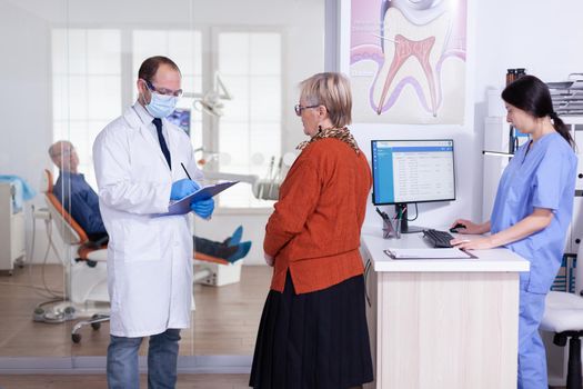 Dentist doctor with face mask explaining diagnosis to senior woman patient in stomatology waiting area hallway. Elderly man sitting on chair for teeth treatment.