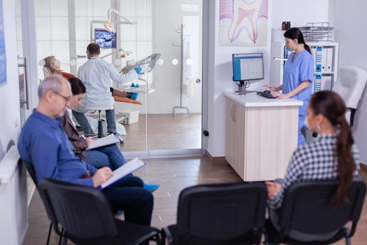 Crowded stomatology waiting area with people filling form for dental consultation. Stomatoloy specialist denstiry treating senior woman cavity. Receptionist working on computer.