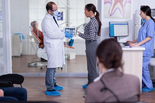 Dentist with face mask pointing at digital teeth radiography x-ray to patient in clinic waiting area. Receptionist writing on computer people appointments.