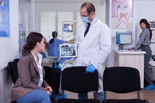 Stomatologist pointing on digital screen explaining x-ray to woman sitting on chair in waiting room of stomatological clinic. Doctor working in modern dental clinic, examining, showing radiography.