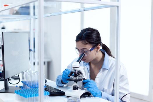 Scientist in laboratory doing research looking through microscope wearing protection glasses and gloves. Chemist wearing lab coat using modern technology during scientific experiment in sterile environment.