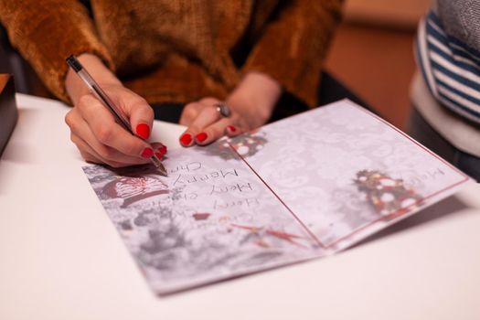 Closeup of woman hands writing xmas greeting card for family during christmas holiday standing in xmas decorated kitchen. Happy family enjoying spending winter season together
