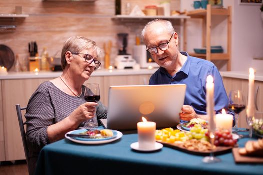 Elderly couple using laptop in the kitchen celebrating relationship. Senior people sitting at the table browsing, searching, using laptop, technology, internet, celebrating their anniversary in the dining room.