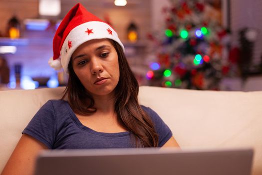 Adult resting on couch reading business email using laptop computer searching information using laptop computer. Caucasian female spending christmastime working. Celebrating winter season