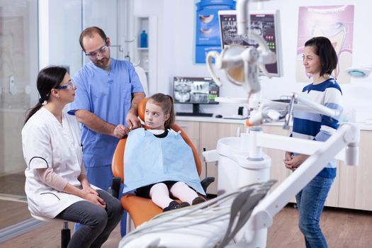 Dentist assistant preparing kid patient for tooth examination in dental office. Child with her mother during teeth check up with stomatolog sitting on chair.