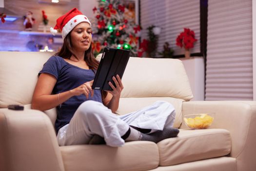 Woman sitting in lotus position on sofa browsing on social media writing xmas email messaging with friends using tablet computer. Adult person celebrating christmas season enjoying winter holiday