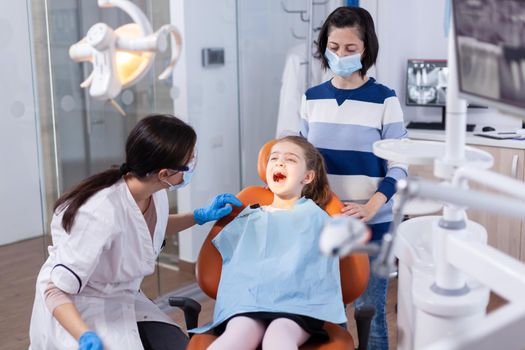 Kid keeping mouth open sitting on dental chair in the course of oral examinantion. Dentistry specialist during child cavity consultation in stomatology office using modern technology.