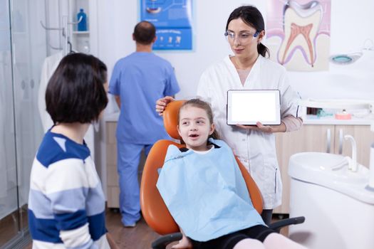 Little girl sitting in dental chair and pediatric dentist using tablet pc with chroma key. Stomatolog explaining teeth prevention to mother and child holding tablet pc with copy space available.