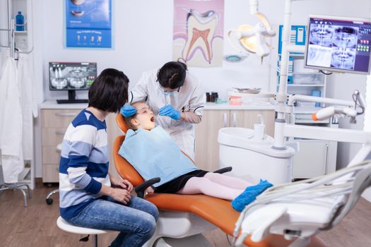 Kid patient in dentistiry office getting treatment for teeth cavity sitting on dental chair wearing bib. Dentistry specialist during child cavity consultation in stomatology office using modern technology.