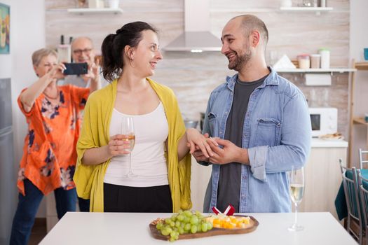 Young man smiling at wife while putting engagement ring on her finger in front of parents during family lunch.