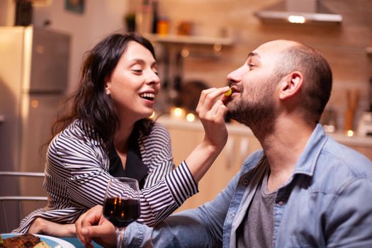 Husband feeding wife with grapes during romantic dinner celebrating relationship. Wife and husband celebrating anniversary with red wine, tender moments at candle lights in kitchen.