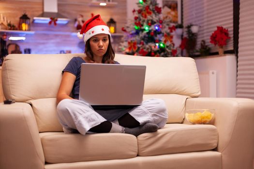 Woman with santa hat resting on couch reading business email using laptop computer searching information using laptop computer. Girl spending christmas time working. Winter season