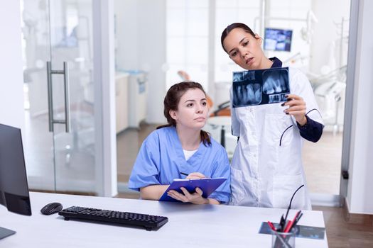 Dentist holding teeth x-ray explaning diagnosis for nurse at reception. Stomatolog and her assistant in reception of dental clinic looking at teeth radiography.