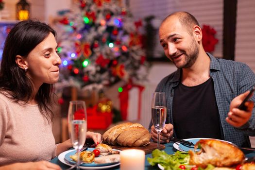 Happy family eating delicious dinner sitting at dining table in xmas decorated kitchen celebrating christmas holiday. Couple watching entertainment movie show using phone. Santa-claus season