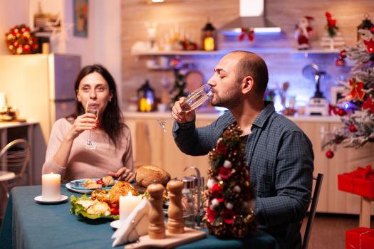 Happy romantic couple drinking wine while sitting at dinning table in xmas decorated kitchen. Cheerful family celebrating christmastime enjoying spending winter holiday together