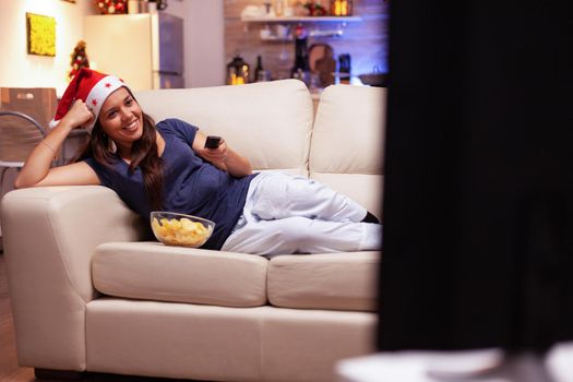 Adult person smiling while watching xmas movie lying on sofa in -xmas decorated kitchen during christmastime. Woman celebrating christmas holiday at home enjoying winter season