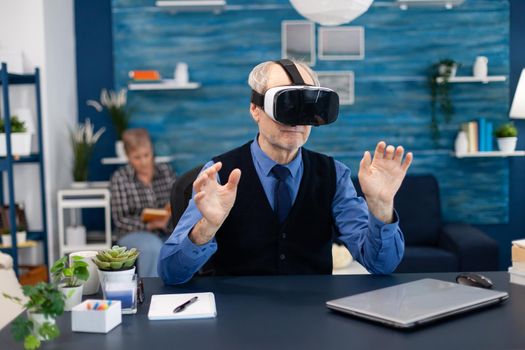 Cheerful old man wearing virtual reality headset sitting at desk office. Elderly man pensioner in living room sitting at desk with futuristic goggles for augmented cyberspace wife watching tv.