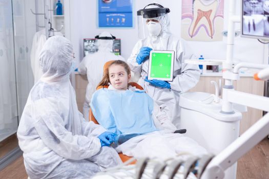 Tablet pc with green screen used by dentist in ppe suit during consultation of little girl. Medical specialist in oral hygine holding tablet pc with copy space available during global pandemic with covid19.