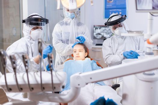 Mother dressed in ppe suit comforting daughter in the course of dental treatment. Stomatology team wearing ppe suit during covid19 doing procedure on child teeth.