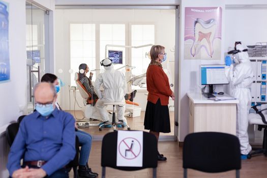 Dentist doctor in ppe suit consulting patient in dental clinic dressed in ppe suit as safety precation during global pandemic with coronaivurs. Senior woman talking with dentistiry stomatology receptionist sitting on chair.