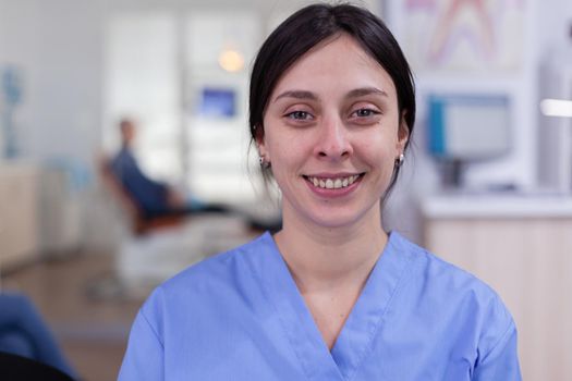 Dental assistant looking at camera while doctor examining patient in background. Professional stomatologist nurse smiling on webcam sitting on chair in waiting room of stomatological clinic.