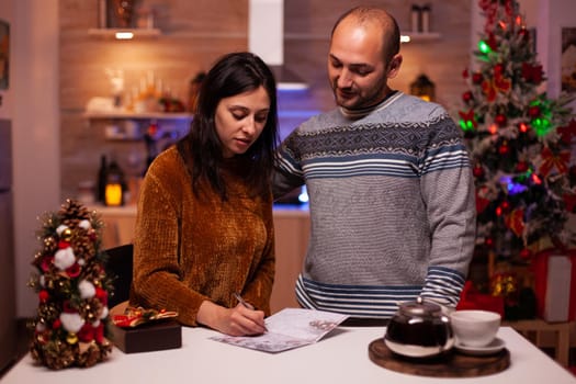 Cheerful girlfriend writing christmas greeting postcard celebrating xmas winter season with boyfriend standing in x-mas decorated kitchen, Happy family enjoying spending christmastime together