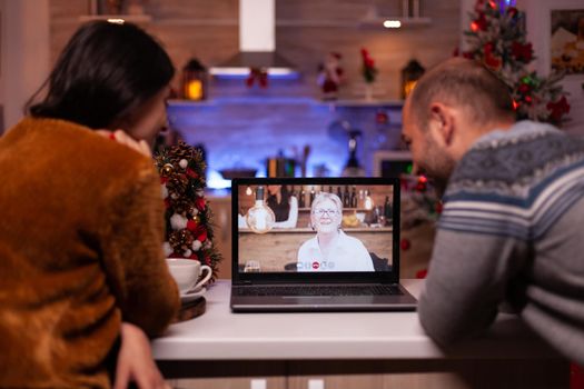 Joyful family talking with grandmother during online videocall meeting on laptop computer celebrating christmastime together in xmas decorated kitchen. Happy couple enjoying winter season