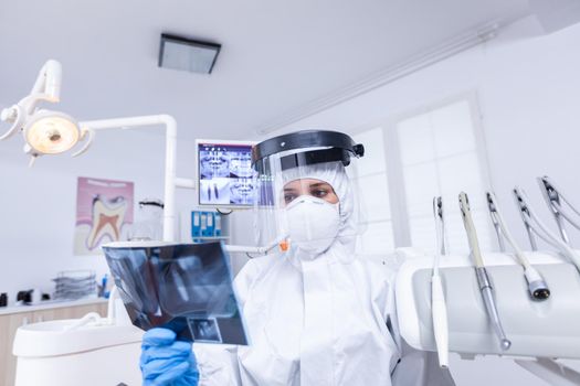 Patient pov in dental office looking at dentist holding radiography dressed in protective gear during covid-19. Dental specialist wearing protective hazmat suit against coroanvirus showing radiography.